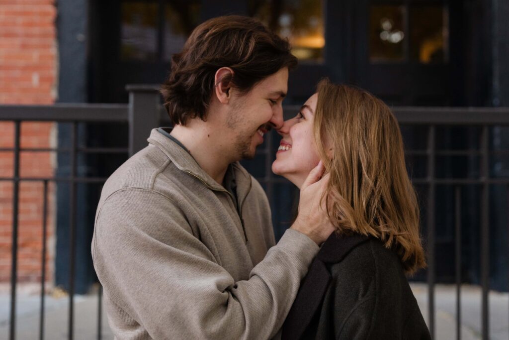 Couple leaning in for a kiss near black iron railings and red brick buildings during a downtown Denver engagement session