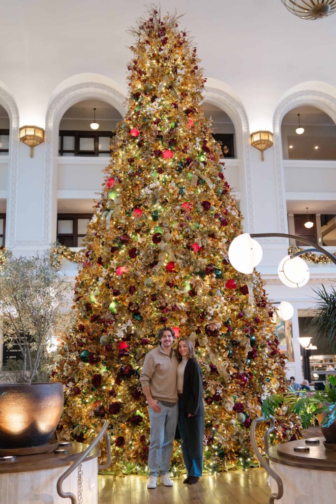 Couple standing in front of a massive Christmas tree inside Denver Union Station during their holiday engagement session