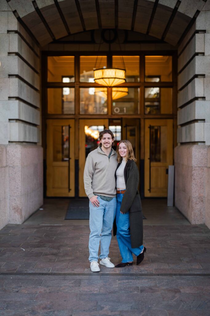 Couple standing together under an arched entrance with warm lighting behind them.