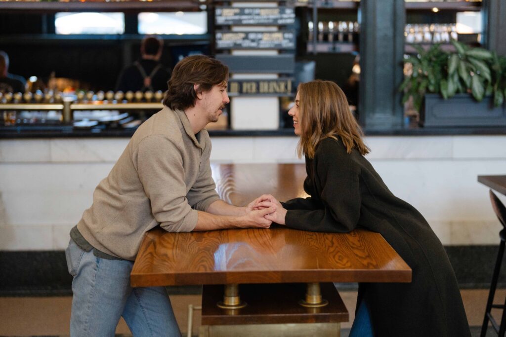 Couple leaning over a wooden table inside Denver Union Station, holding hands and smiling at each other.