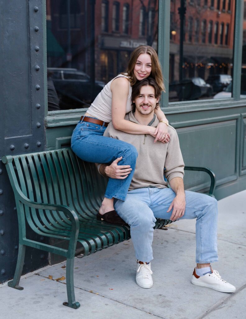 Couple sitting on a green bench in downtown Denver, the woman wrapped around her partner’s shoulders during their engagement session.