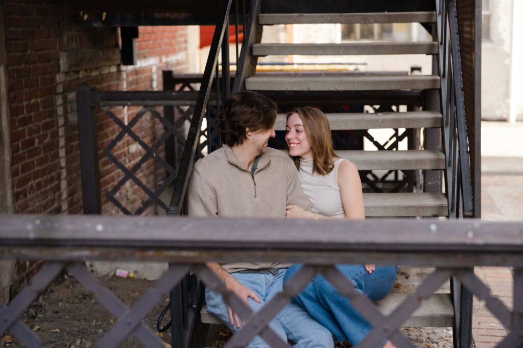 Couple sitting together on metal stairs in an alley, leaning in close and smiling warmly.