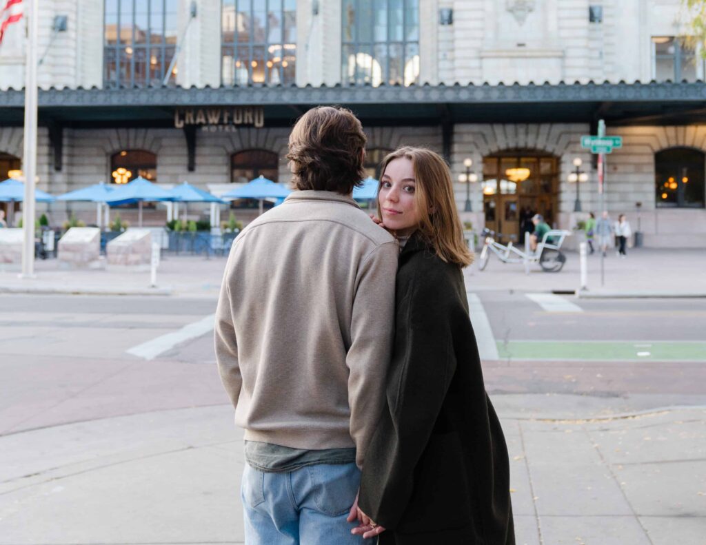 She looks back over her shoulder while holding hands in front of Union Station during their Denver Union Station engagement session